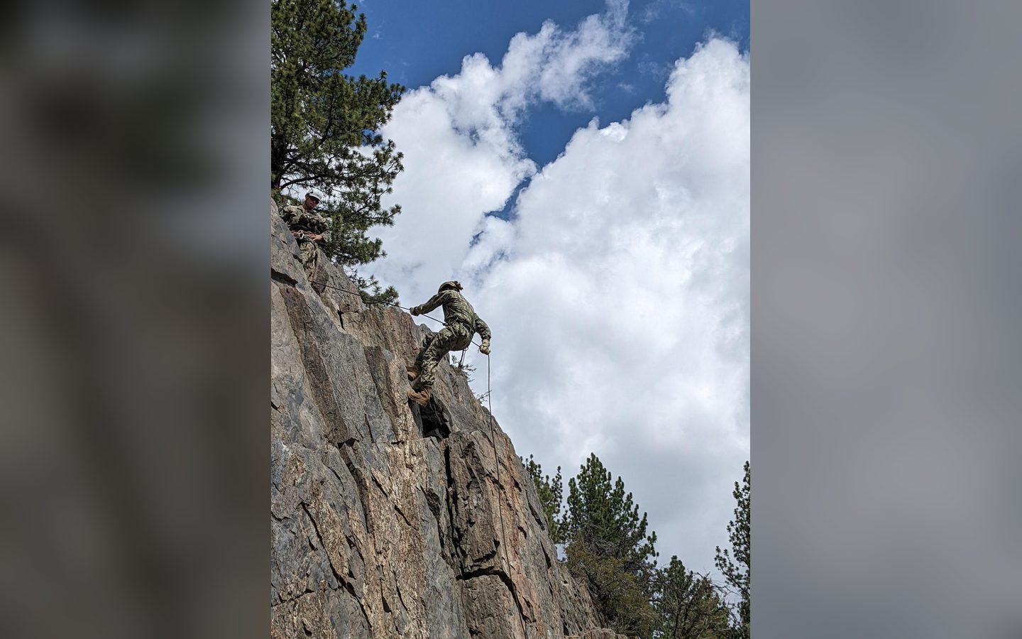 A climber on a rope halfway up a rock face under summer clouds