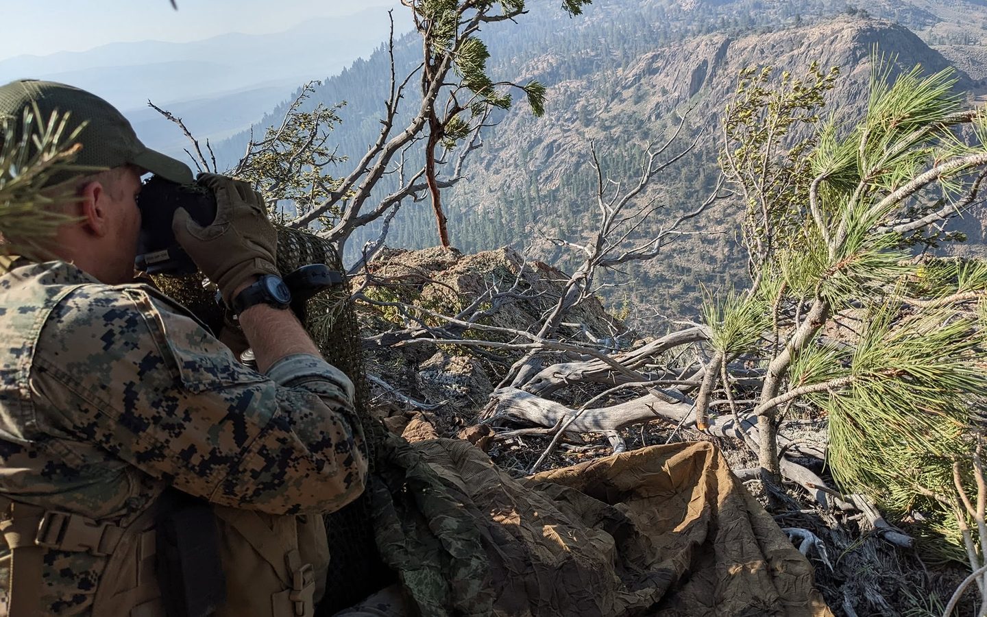A Marine glassing a mountain ridge through binoculars from a hide