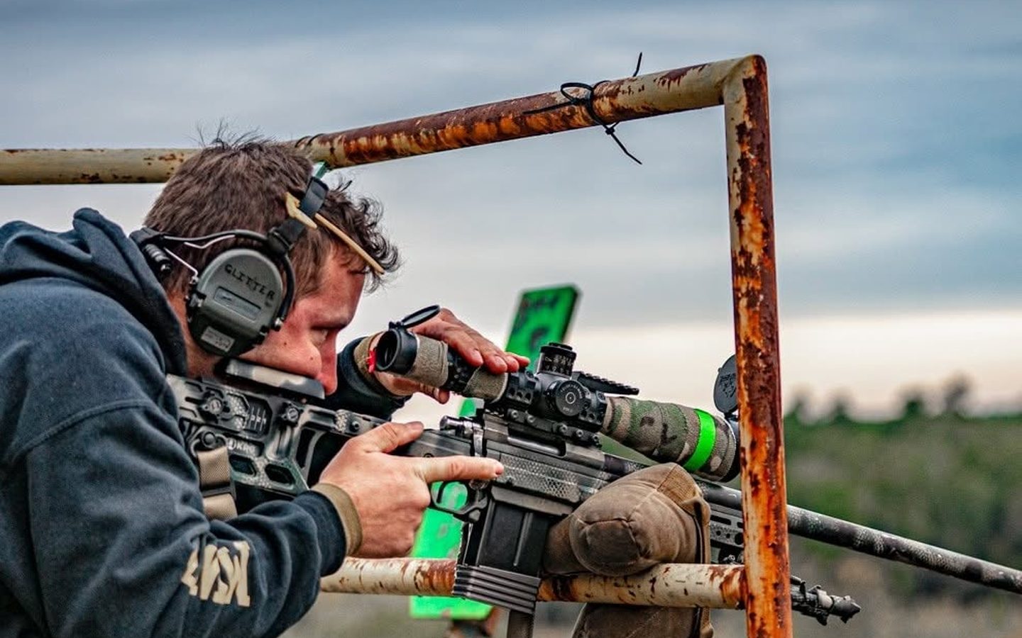 A shooter on a rusted barricade, rifle braced on a bag, glassing through the scope