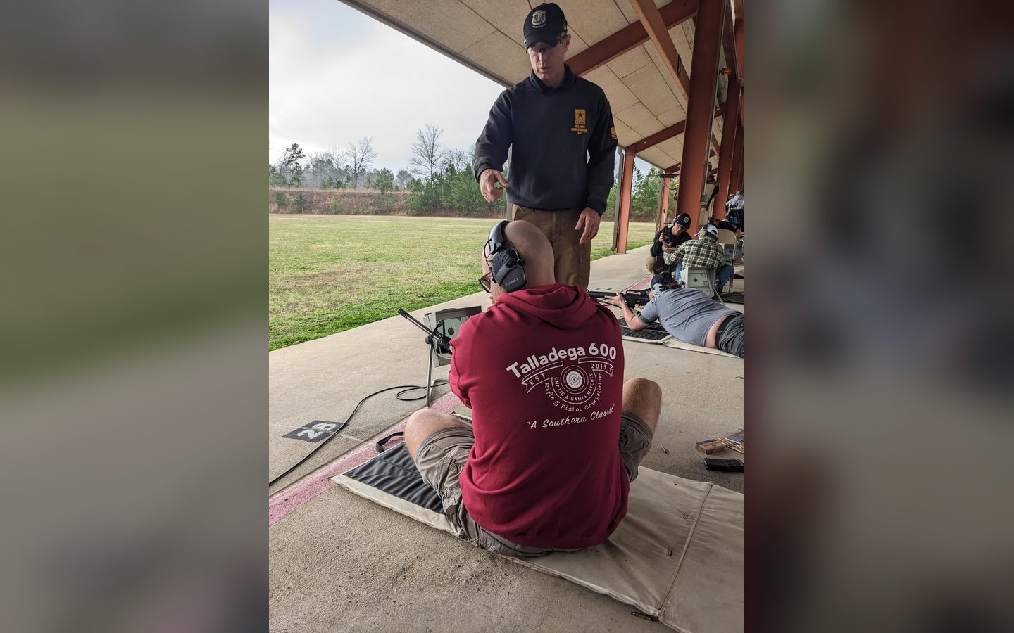 A shooter seated on the firing line under a range cover, a coach standing behind