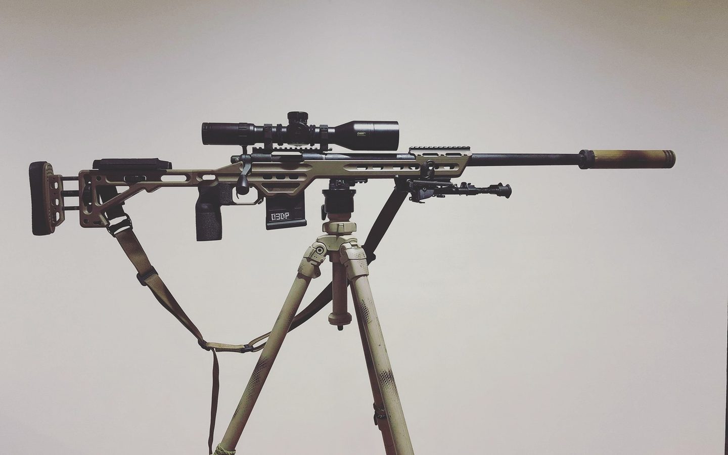 A precision rifle with a suppressor and scope on a camouflaged tripod